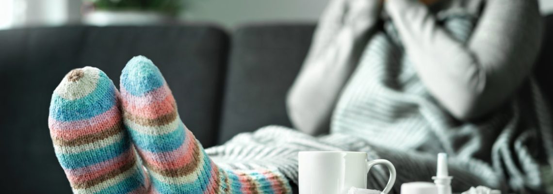 Sick woman with flu, cold, fever and cough sitting on couch at home. Ill person blowing nose and sneezing with tissue and handkerchief. Woolen socks and medicine.