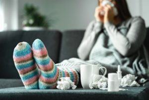 Sick woman with flu, cold, fever and cough sitting on couch at home. Ill person blowing nose and sneezing with tissue and handkerchief. Woolen socks and medicine.